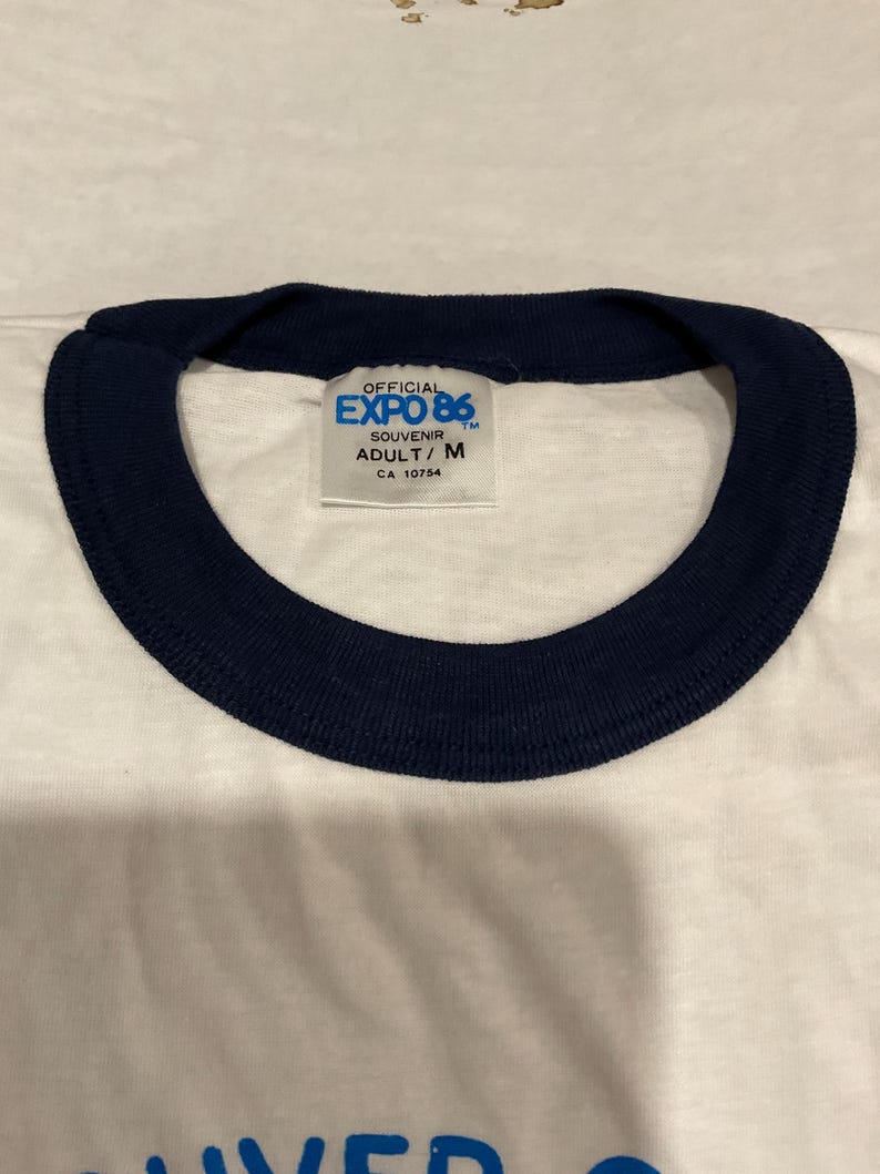 Puede incluir: Camiseta blanca con cuello azul marino. La camiseta presenta el texto "OFFICIAL EXPO 86" en una etiqueta. La etiqueta tambi&eacute;n incluye el texto "SOUVENIR ADULT/ M CA 10754".