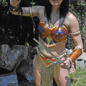 May include: A woman wearing a gold and blue costume with a gold chain skirt and a headpiece. She is holding a silver weapon with a curved blade. The woman is standing in a woodland setting.