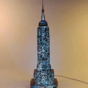 May include: A tall, dark, architectural model of the Empire State Building featuring a grid of illuminated blue windows. The structure has a stepped design with a central spire at the top, emitting a bright blue light from within the building frame.