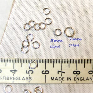 May include: A collection of silver-coloured metal jump rings in two sizes. Text labels indicate 5mm (20ga) and 7mm (18ga) sizes. A ruler is visible at the bottom, showing measurements in inches, with the text MADE IN ENGLAND printed on it.