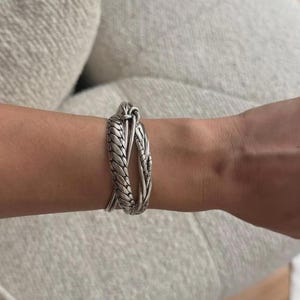 May include: A silver-toned metal cuff bracelet featuring a twisted, rope-like texture. The design consists of two overlapping strands that meet at a central point, creating an open, layered appearance that wraps around the wrist.