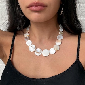 Op de afbeelding: Een ketting bestaande uit een enkele streng van ronde, iriserende witte schelpenschijfjes van verschillende groottes. De schijfjes zijn in een doorlopende rij gerangschikt en vertonen natuurlijke parelmoertexturen en subtiele crèmekleurige tinten.