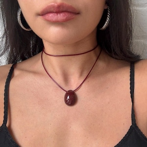 Puede incluir: Un colgante ovalado de color rojo oscuro en un collar de cordón fino a juego, usado como gargantilla con una capa de cordón adicional. El colgante tiene un acabado liso y pulido. Se ven pendientes de aro trenzados plateados a ambos lados.