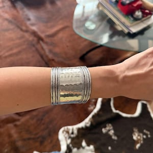 May include: A wide silver cuff bracelet with a hammered texture and detailed edges. The bracelet is worn on a person's arm, showcasing its intricate design and metallic sheen. The cuff is approximately 2 inches wide.