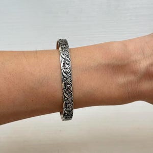 May include: A silver cuff bracelet with an engraved swirling pattern. The bracelet is worn on a wrist and has a polished finish. The design features a repeating motif of curved lines and spirals, creating a textured look. The jewelry is a fashion accessory.