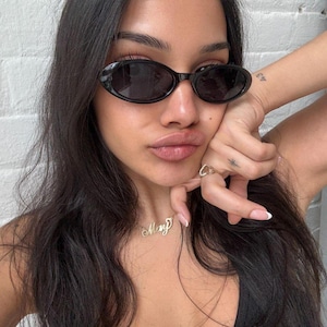 Könnte beinhalten: Schwarze ovale Sonnenbrille mit schmalem Rahmen. Eine goldene Halskette mit dem Namen "Mary" in Schreibschrift. Ein goldener Ring und ein kleines Tattoo sind ebenfalls sichtbar.
