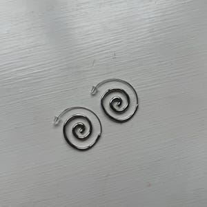 Puede incluir: Un par de pendientes en espiral de color plateado. Cada pendiente presenta un diseño en espiral con un poste para orejas perforadas. Los pendientes se muestran sobre una superficie de madera blanca.