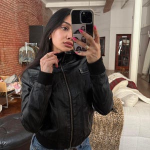 May include: Black leather jacket with a ribbed collar and cuffs. The jacket has a zipper closure and two small pockets. The person in the image is wearing blue jeans.