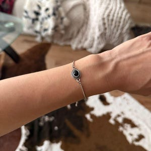 Könnte beinhalten: Ein silbernes Armband mit einem kleinen, runden, schwarzen Edelstein in der Mitte. Der Edelstein ist in einer dekorativen, gemusterten Lünette gefasst. Das Armband ist zierlich und wird am Handgelenk getragen.