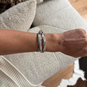Op de afbeelding: Een zilverkleurige armband met een slangenontwerp gewikkeld rond een pols. De armband heeft een textuur, schubbenachtig patroon en een slangenkopsluiting. De sieraden worden gedragen op een blanke arm.