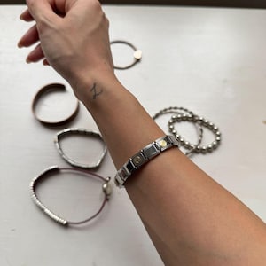 Puede incluir: Una colección de pulseras de varios estilos. Una pulsera de eslabones plateados con detalles dorados se lleva en la muñeca. Otras pulseras incluyen una pulsera de cuentas plateadas, un brazalete marrón y una pulsera plateada con un cordón rojo.