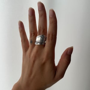 Puede incluir: Un anillo plateado con intrincados detalles florales y de volutas. El anillo está hecho de cubiertos reciclados, mostrando una estética vintage única. El anillo se lleva en una mano sobre un fondo blanco.