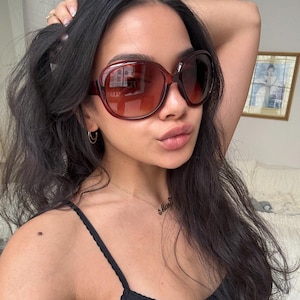 May include: Oversized, dark red sunglasses with brown gradient lenses. The sunglasses have a thick frame and are worn by a person with dark hair. The image is a close-up, showing the sunglasses and part of the person's face.