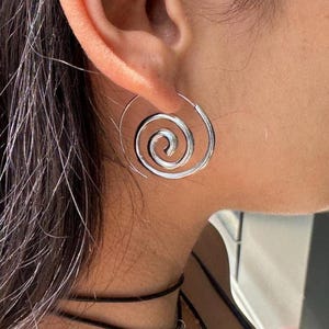 May include: A silver-toned spiral hoop earring featuring a circular, coiled wire design that loops inwards. The earring is constructed from a polished metallic material and is worn through the earlobe, showcasing a continuous, smooth, and rounded shape.