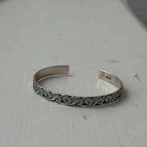 Pode incluir: Uma pulseira de punho de prata com um padrão de ondas repetidas. A pulseira aberta é marcada com "925" no interior. A pulseira tem aproximadamente 0,6 cm de largura.