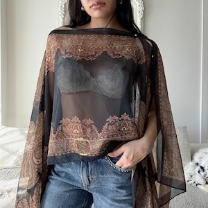 May include: A sheer black cape top with a paisley pattern in shades of brown and gold. The top has a wide neckline and a decorative gold buckle. The sleeves are open, creating a flowing silhouette. The top is paired with blue jeans.