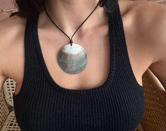 May include: A large, round pendant necklace with a natural, iridescent shell design. The pendant is suspended from a black cord. The person is wearing a black ribbed tank top.