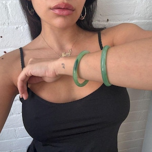 May include: Two jade green bangles are worn on an arm, with a black tank top in the background. A gold necklace with the name "May" is visible, along with small gold hoop earrings. A tattoo of the numbers "43" is on the wrist.