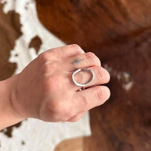 May include: A silver-colored ring with an oval design. The ring is worn on a finger. The ring is a simple, modern piece of jewelry.