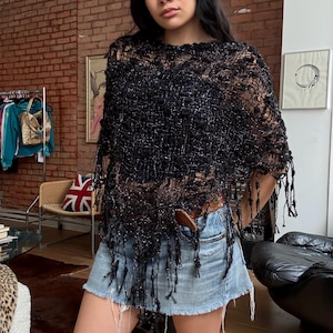 May include: A black, loose-knit, semi-sheer poncho featuring a metallic shimmer, textured yarn accents, and long fringe detailing along the hemline. The garment has a wide, open-weave construction and is worn over a simple dark base layer.