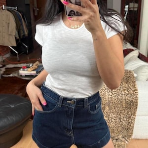 May include: A white crew neck t-shirt paired with dark blue denim shorts. The shorts have a classic five-pocket design. The outfit is simple and casual.