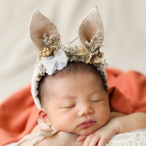 May include: A newborn baby wearing a beige crochet headband with two upright felt bunny ears, decorated with small dried flowers and a white lace bow. The baby is resting on a white lace-trimmed cushion inside a basket with a rust-coloured fabric.