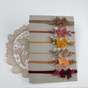 Peut inclure: Un ensemble de six bandeaux élastiques fins disposés verticalement sur une carte rectangulaire. Chaque bandeau présente un petit nœud en tissu centré sur un bouquet d'éléments floraux séchés dans des tons beige, vieux rose, jaune moutarde, sable, orange et bordeaux.