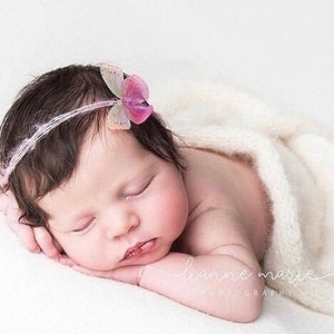 May include: A newborn baby with dark hair is peacefully sleeping, wrapped in a soft, white blanket. The baby wears a delicate pink headband with a butterfly accent. The image is a professional newborn portrait.