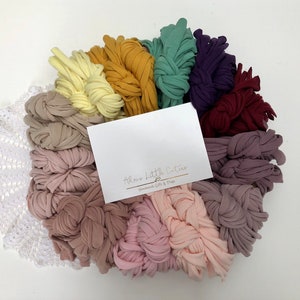 May include: A variety of colors of soft, stretchy fabric yarn arranged in a circle. The yarn is in shades of yellow, orange, green, purple, red, pink, and brown. The yarn is arranged in a circle with a white crocheted doily in the center. A white card with the text "Almost Little Cuties Handmade Gifts & Props" is in the center of the yarn.