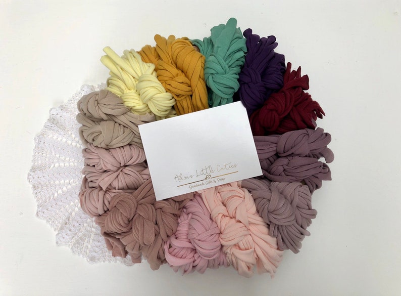 May include: A variety of colors of soft, stretchy fabric yarn arranged in a circle. The yarn is in shades of yellow, orange, green, purple, red, pink, and brown. The yarn is arranged in a circle with a white crocheted doily in the center. A white card with the text "Almost Little Cuties Handmade Gifts & Props" is in the center of the yarn.