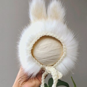May include: A white bonnet featuring long, plush faux fur trim around the face opening and two upright, pointed faux fur ears on top. The base of the bonnet is constructed from a cream-colored crocheted yarn, complete with matching braided ties.