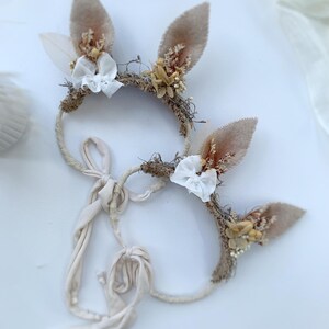 May include: Two headbands featuring beige felt animal ears adorned with dried flowers, small white fabric bows, and textured natural fibers. Each headband includes long, cream-colored fabric ties for securing the accessory in place.