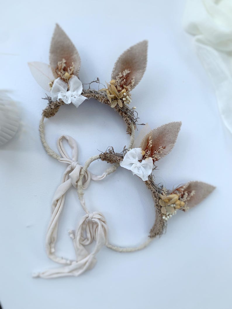 May include: Two headbands featuring beige felt animal ears adorned with dried flowers, small white fabric bows, and textured natural fibers. Each headband includes long, cream-colored fabric ties for securing the accessory in place.
