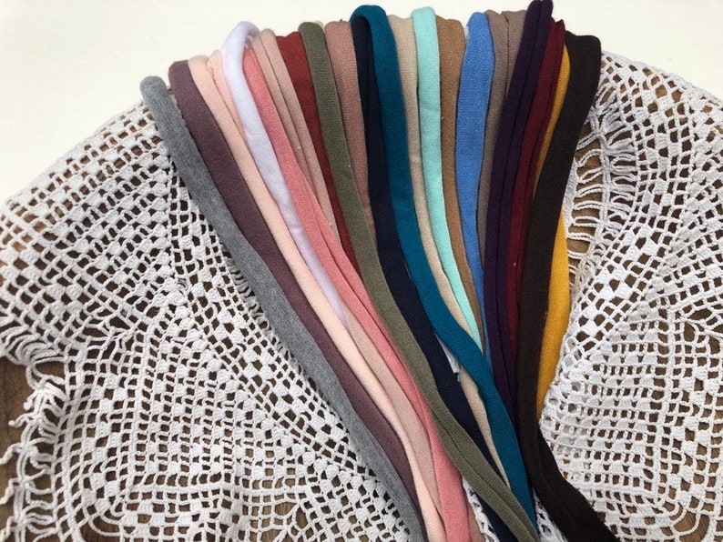 Puede incluir: Una colecci&oacute;n de 20 diademas de tela de diferentes colores, dispuestas en forma de abanico sobre una superficie de crochet blanco.