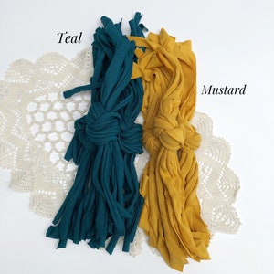 May include: Two bundles of fabric yarn, one teal and one mustard yellow, are arranged on a white surface with a white crocheted doily underneath. The fabric yarn is made from recycled t-shirts.