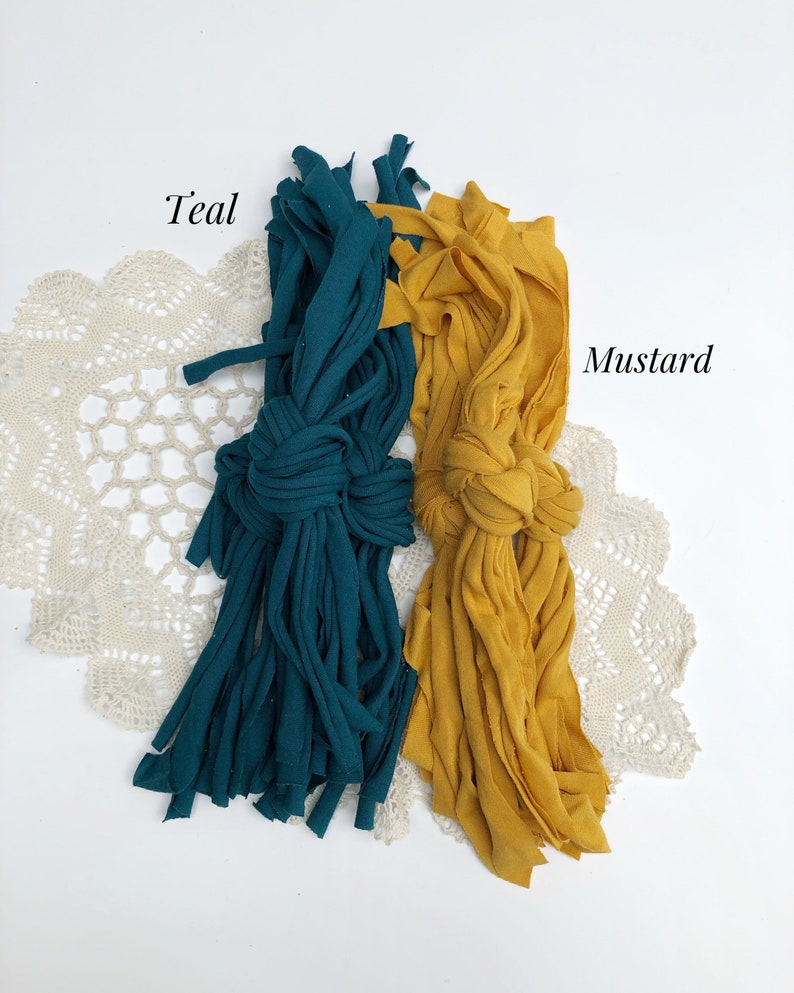 May include: Two bundles of fabric yarn, one teal and one mustard yellow, are arranged on a white surface with a white crocheted doily underneath. The fabric yarn is made from recycled t-shirts.