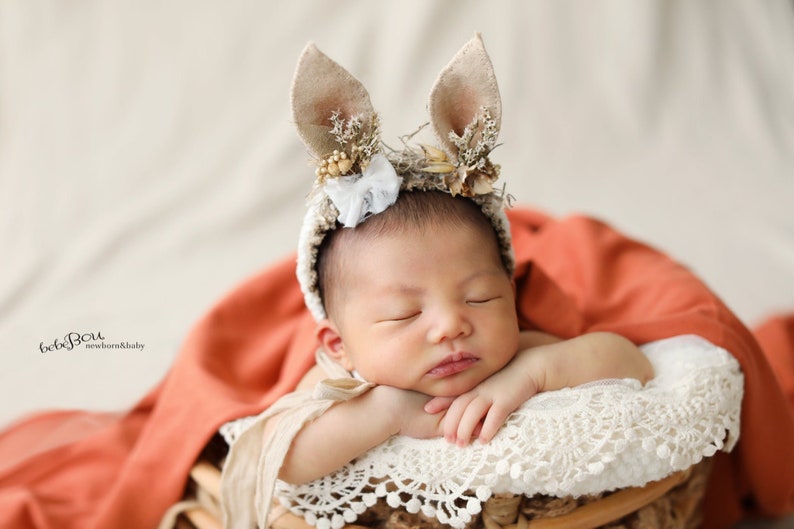 May include: A newborn baby wearing a beige crochet headband with two upright felt bunny ears, decorated with small dried flowers and a white lace bow. The baby is resting on a white lace-trimmed cushion inside a basket with a rust-coloured fabric.