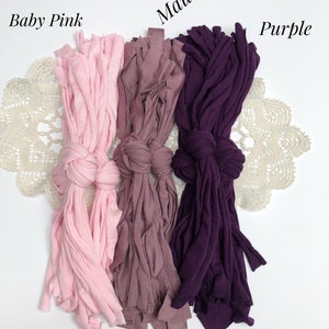 May include: Three bundles of fabric yarn in different colors: baby pink, mauve, and purple. The yarn is arranged in a row on a white surface with a lace doily underneath.