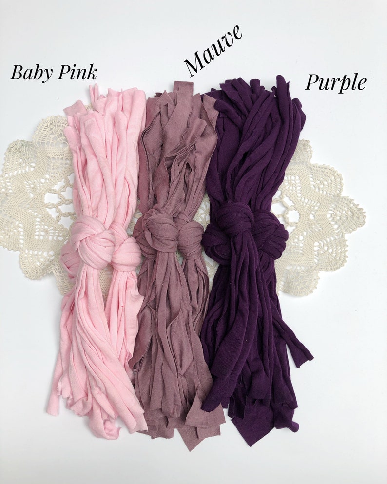 May include: Three bundles of fabric yarn in different colors: baby pink, mauve, and purple. The yarn is arranged in a row on a white surface with a lace doily underneath.
