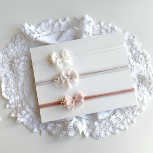 Può includere: Tre fasce elastiche sottili disposte orizzontalmente su un cartoncino rettangolare bianco. Ogni fascia presenta un piccolo fiocco in tessuto bianco, beige chiaro o rosa polvere, decorato con minuscoli rametti di fiori bianchi sul lato.