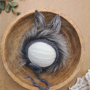 May include: A gray faux fur bonnet featuring two pointed ears on top and a crocheted dark gray trim around the face opening. The bonnet includes a matching crocheted chin strap with a small decorative flower detail attached to the side.