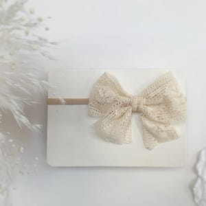 May include: A cream-coloured lace bow attached to a thin, matching elastic headband. The bow features an intricate floral pattern and is displayed on a rectangular white card, showcasing the delicate texture and soft neutral tone of the fabric.