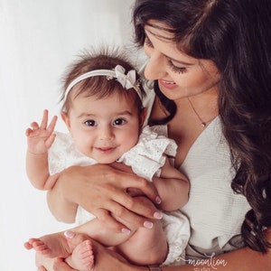 K&ouml;nnte beinhalten: Eine Frau h&auml;lt ein Baby M&auml;dchen, das ein wei&szlig;es Kleid mit Spitzenbesatz tr&auml;gt. Das Baby l&auml;chelt und tr&auml;gt ein wei&szlig;es Stirnband mit einer Schleife auf dem Kopf. Die Frau tr&auml;gt ein wei&szlig;es Oberteil und hat lange dunkle Haare.