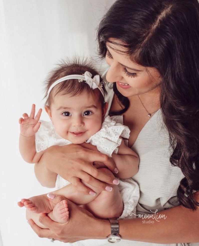 K&ouml;nnte beinhalten: Eine Frau h&auml;lt ein Baby M&auml;dchen, das ein wei&szlig;es Kleid mit Spitzenbesatz tr&auml;gt. Das Baby l&auml;chelt und tr&auml;gt ein wei&szlig;es Stirnband mit einer Schleife auf dem Kopf. Die Frau tr&auml;gt ein wei&szlig;es Oberteil und hat lange dunkle Haare.