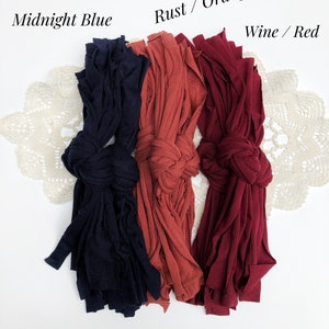 May include: Three bundles of fabric yarn in different colors: midnight blue, rust orange, and wine red. The yarn is arranged in a row on a white surface.