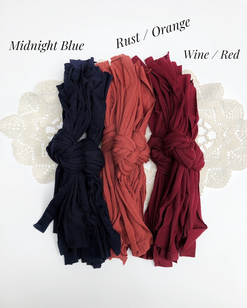 May include: Three bundles of fabric yarn in different colors: midnight blue, rust orange, and wine red. The yarn is arranged in a row on a white surface.