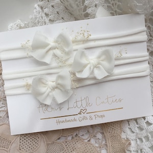 K&ouml;nnte beinhalten: Drei wei&szlig;e Stoffhaarb&auml;nder mit einem wei&szlig;en elastischen Haarreifen. Das Haarreifen befindet sich auf einer wei&szlig;en Karte mit dem Text "Adore Little Cuties Handmade Gifts & Props".