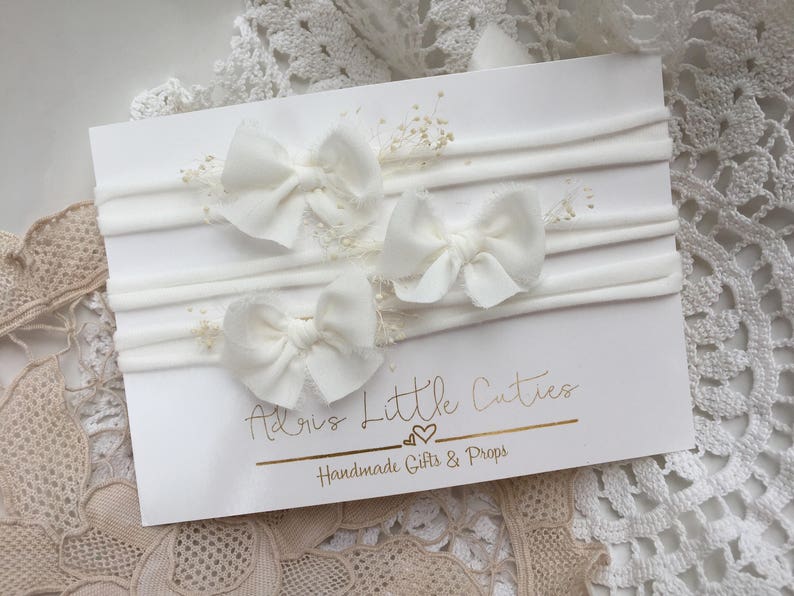 K&ouml;nnte beinhalten: Drei wei&szlig;e Stoffhaarb&auml;nder mit einem wei&szlig;en elastischen Haarreifen. Das Haarreifen befindet sich auf einer wei&szlig;en Karte mit dem Text "Adore Little Cuties Handmade Gifts & Props".