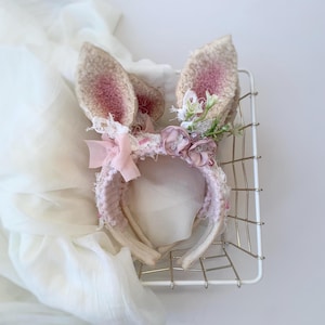 May include: A headband featuring two upright, textured, beige rabbit ears with pink-tinted centers. The band is decorated with a small pink ribbon bow, two dusty rose fabric flowers, and delicate white lace and green foliage accents.