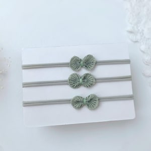 May include: Three sage green elastic headbands, each featuring a small, textured, fan-shaped bow detail at the center. The headbands are displayed horizontally on a plain white rectangular card, showcasing a consistent design and color palette.